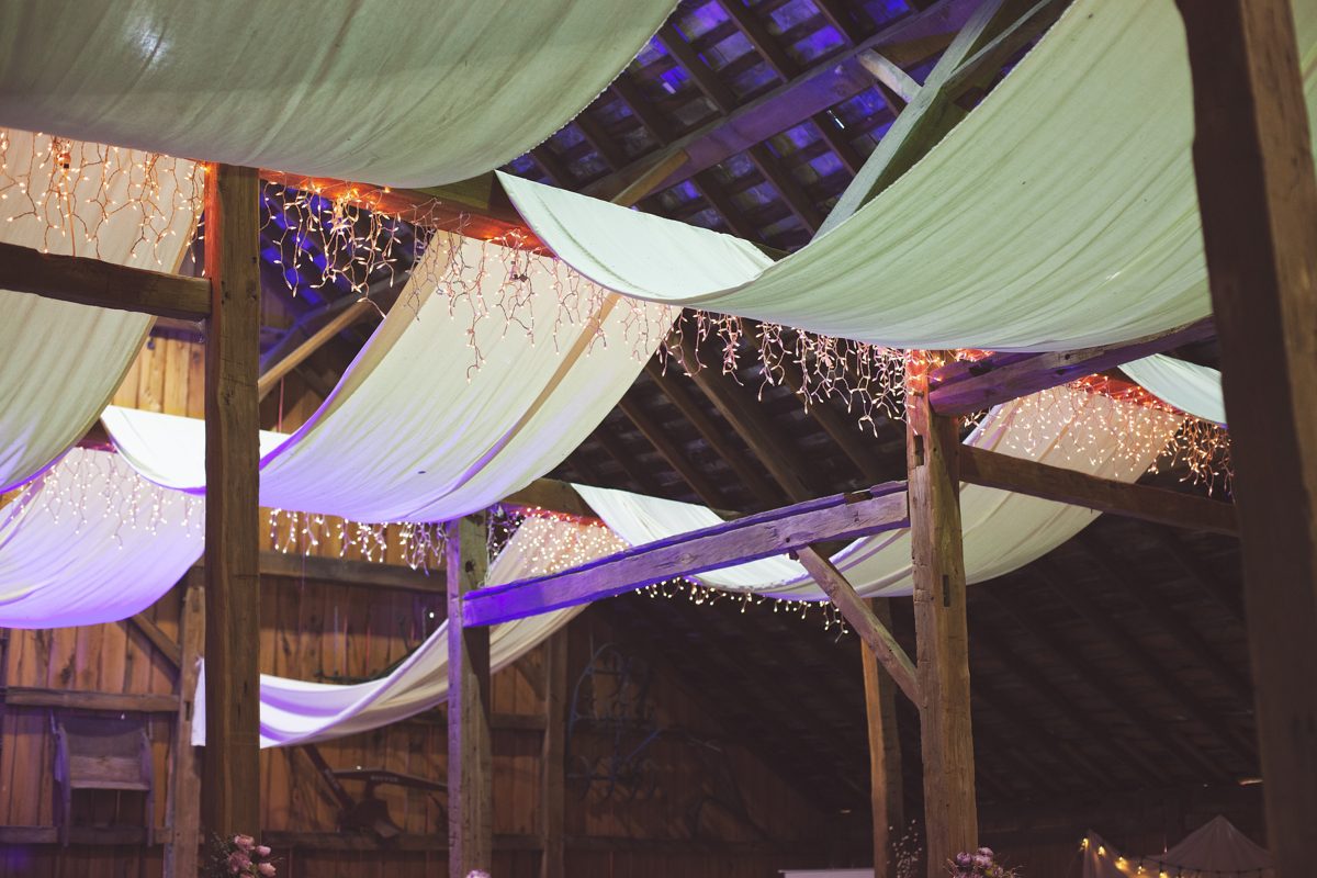 White fabric and string lights draped across the beams of the barn for a wedding