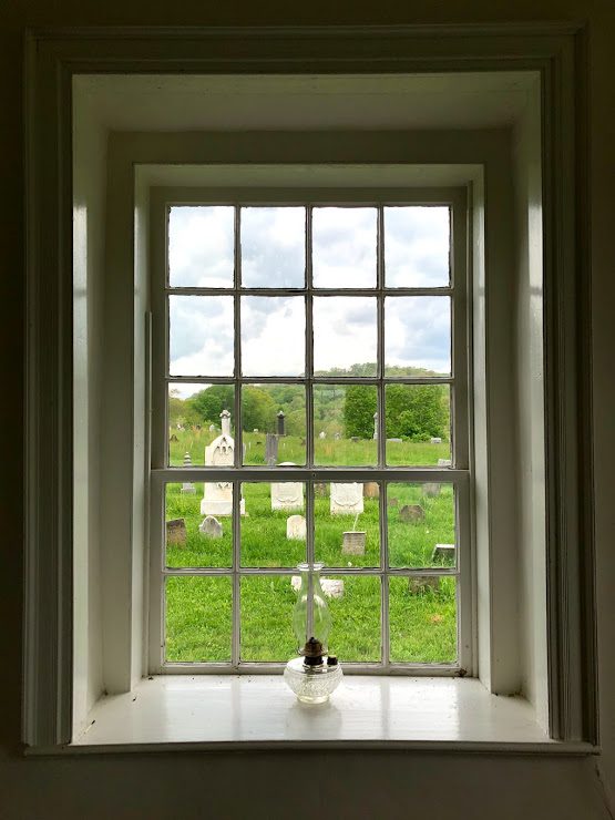 A deep-set window in the meetinghouse with an oil lamp on the sill and the cemetery beyond